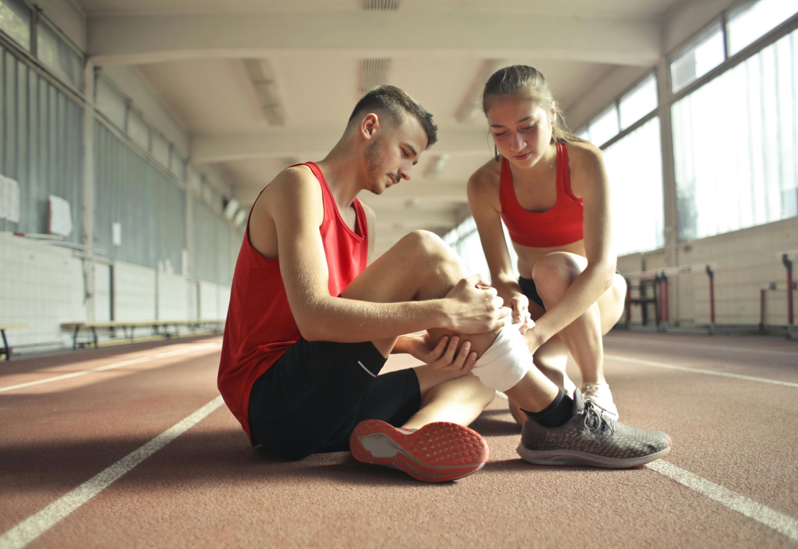 Two athletes on an indoor track, one experiencing knee pain and the other assisting